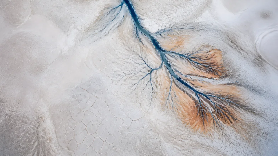 A cold blue channel branches through alabaster salt and restrained copper sediment fans.